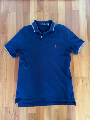 Marinblå pikétröja från Polo Ralph Lauren - Klassisk marinblå pikétröja från Polo Ralph Lauren med röd broderad logga på bröstet. Tröjan har korta ärmar, krage med röd och vit rand samt knappar vid halsen. Perfekt till jeans eller shorts för en avslappnad men snygg look. Säljes då den har blivit för liten, endast använd 4 gånger ungefär!