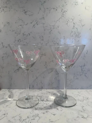 Två cocktailglas med rosa rosetter - Säljer två genomskinliga cocktailglas med tunna ben och breda kupor. Glasen är dekorerade med små rosa rosetter runt om, vilket ger dem en söt och lekfull stil. Perfekta för drinkar eller som dekoration i köket.