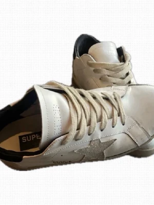 Vita Golden Goose  - Snygga vita sneakers från Golden Goose med grå mockastjärna på sidan och svarta detaljer vid hälen. Skorna har beige snörning och klassisk låg modell i läder. 