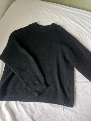 Svart stickad tröja från H&M - En svart stickad tröja från H&M med rund halsringning och långa ärmar. Tröjan har en enkel och avslappnad passform med ribbstickade muddar vid ärmslut och nederkant. Perfekt för lager-på-lager och en stilren look. Lite nopprig som man kan se på bilderna. 