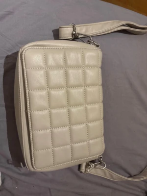 Beige quiltad axelväska i konstläder - Snygg beige axelväska med quiltat mönster och justerbar axelrem. Väskan har flera fack inuti, inklusive ett med dragkedja, och stängs med magnetknapp. Perfekt för dig som gillar stilrena accessoarer i konstläder.