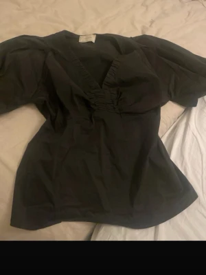 Svart puffärmblus från Neo Noir - Trendig svart blus från Neo Noir med djup v-ringning och puffiga korta ärmar. Blusen har en figurnära passform och lätt glansigt tyg som ger en snygg look. Perfekt att styla med både jeans och kjol.