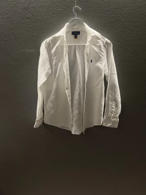 Vit linneskjorta från Polo Ralph Lauren - Klassisk vit skjorta från Polo Ralph Lauren i luftigt linne. Skjortan har lång ärm, knappar framtill och den ikoniska broderade loggan på bröstet. Perfekt för en clean och stilren look.
