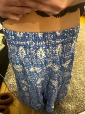 Blå långkjol med mönster - Superskön blå långkjol med vitt mönster och smock i midjan. Kjolen har en avslappnad passform och är perfekt för varma dagar. Tillverkad i ett lätt och luftigt material som känns bekvämt hela dagen.