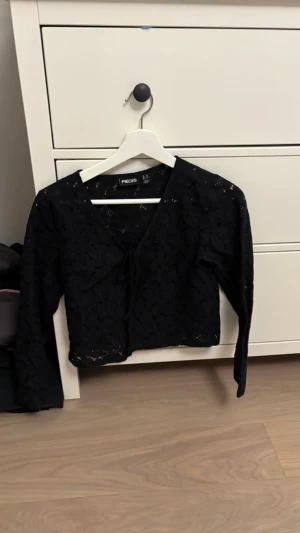 Svart spetsig topp från Pieces - Snygg svart långärmad topp från Pieces i genombruten spets med knytning framtill. Toppen har v-ringning och är figurnära, perfekt för att lyfta en outfit med lite extra edge.