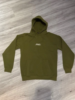 Olivgrön hoodie från Daily Essentials - Snygg olivgrön hoodie från Daily Essentials med vit logga på bröstet och stort tryck på ryggen. Klassisk känguruficka framtill, ribbade muddar och justerbar huva. Storlek M
