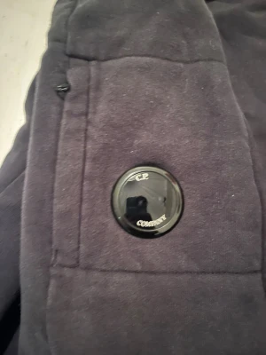 Svarta mjukisbyxor från C.P. Company - Säljer ett par svarta mjukisbyxor från C.P. Company med den ikoniska linsen på benfickan. Byxorna har resår i midjan och mudd vid bensluten. Tillverkade i mjukt och bekvämt material som passar perfekt för en avslappnad stil.