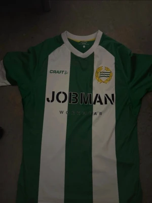 Hammarby matchtröja Craft grön/vit - Säljer en Hammarby matchtröja från Craft i storlek M. Tröjan har gröna och vita vertikala ränder, klubbmärke på bröstet och sponsorlogga 'JOBMAN WORKWEAR' framtill. Tillverkad i lätt och ventilerande material, perfekt för fotboll eller träning.