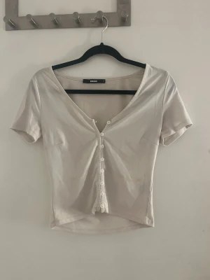 Beige croppad topp från Bik Bok - Snygg vit croppad topp från Bik Bok med v-ringning och korta ärmar. Toppen har knappar framtill och är tillverkad i mjuk bomull som känns skön mot huden. Perfekt till jeans eller kjol för en enkel och trendig look.
