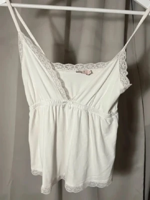 Vit spets kantad linne topp från Nelly!  - Supersöt vit linnetopp från Nelly i storlek XS. Toppen har tunna axelband, v-ringning och är dekorerad med spetsdetaljer längs halsringning och nederkant. Mjuk och lätt bomullskänsla, perfekt för varma dagar. Snyggt rynkad under bysten för en feminin siluett. Buda gärna❤️är ni inte trevliga så tillåter jag tyvärr inte köp🫶OBS! Har sytt upp banden på den!! 