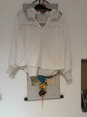 Vit blus med spets från Holly & Whyte - Säljer en vit blus från Holly & Whyte by Lindex med snygga spetsdetaljer på axlar och ärmslut. Blusen har v-ringning, volangkant vid halsen och långa ärmar med breda smockade muddar. Perfekt för en trendig och romantisk look.