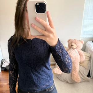 Mörkblå spetsblus med lång ärm - Säljer en mörkblå, långärmad spetsblus med rund hals. Blusen är figurnära och har genombrutna spetsdetaljer över hela plagget. Perfekt att styla med både jeans och kjol för en trendig look.