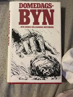 Domedagsbyn och andra sällsamma historier - Samling med sällsamma och kusliga noveller av författare som Roald Dahl, Agatha Christie och William Faulkner. Boken har hårda vita pärmar med röd text och svartvit tecknad illustration på framsidan. Perfekt för dig som gillar spänning och mystik.