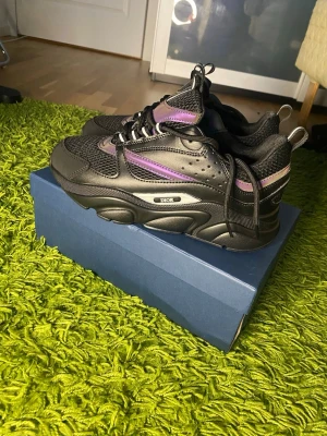 Dior b22 - Chunky svarta Dior sneakers med lila och vita detaljer. Skorna har en blandning av mesh och läder, samt en kraftig sula och reflekterande inslag. Snygg design med sportig vibe och tydlig Dior-logga på sidan. Perfekta för dig som vill sticka ut med exklusiva sneakers.