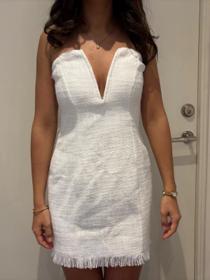 Vit axelbandslös kort klänning - Snygg vit axelbandslös klänning med djup v-ringning och fransdetalj nertill. Klänningen är figurnära och har en strukturerad yta, perfekt för dig som vill sticka ut. Dragkedja bak för enkel påklädning. Perfekt till studenten eller sommarens andra festligheter 