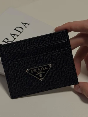 Svart kortfodral från Prada - Snyggt svart kortfodral från Prada i läder med klassisk triangelformad metallogo framtill. Fodralet har flera kortfack och stilren design som passar perfekt i fickan. 