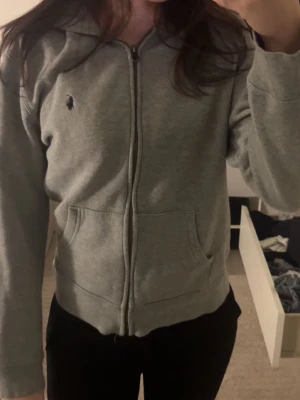 Grå hoodie från Ralph Lauren - Grå zip-hoodie från Ralph Lauren med klassisk huva och två fickor framtill. Liten broderad logga på bröstet i mörkblått. Mjuk bomullskänsla och enkel, stilren design som passar till det mesta.