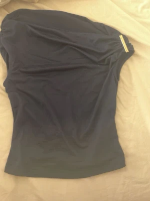 Mörkblå oneshoulder topp med detalj - Snygg mörkblå oneshoulder topp med guldfärgad detalj vid axeln. Tunn och mjuk, perfekt till sommarens outfits. Enkel och stilren design som passar till mycket.