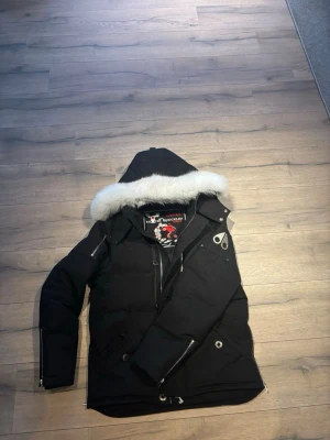 Svart dunjacka med pälskant från Moose Knuckles - Snygg svart dunjacka från Moose Knuckles med stor huva och vit pälskant. Jackan har silverfärgade dragkedjor, logga på ärmen och flera fickor. Perfekt för kalla vinterdagar och riktigt varm tack vare dunfyllningen.
