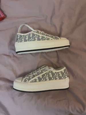 Dior Walk’N Dam Sneakers Skor - Snygga låga sneakers från Dior med gråvitt monogrammönster på canvas och vita snören. Skorna har en tjock vit sula med svart rand och klassisk siluett. Perfekta för dig som gillar streetstyle och vill ha något trendigt och stilrent.