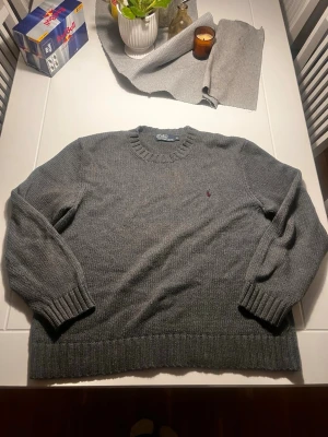 Grå stickad tröja från Polo Ralph Lauren - Säljer en klassisk grå stickad tröja från Polo Ralph Lauren med rund halsringning och ribbade muddar. Tröjan har den ikoniska röda loggan broderad på bröstet och är perfekt för lager på lager. Materialet är mjukt och håller värmen.