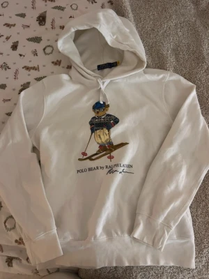 Vit hoodie Polo Bear Ralph Lauren - Vit hoodie från Ralph Lauren med ikoniska Polo Bear-motivet på framsidan där björnen åker skidor. Klassisk huva med dragsko och ribbade muddar. Mjuk bomullskänsla och avslappnad passform, perfekt för en chill och stilren look.