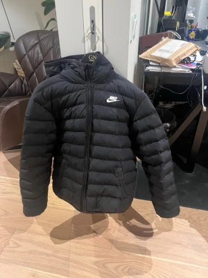 Svart pufferjacka med huva från Nike - Snygg svart pufferjacka från Nike med huva och dragkedja framtill. Jackan har quiltad design, två sidofickor och Nike-logga på bröstet. Perfekt för kalla dagar och enkel att matcha med allt. Klassisk passform och riktigt skön att bära.