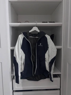 Svartvit vindjacka från Peak Performance - Snygg vindjacka från Peak Performance i svart och vitt. Jackan har huva, dragkedja framtill och två sidofickor med dragkedja. Perfekt för blåsiga dagar och sportiga outfits. Materialet är lätt och smidigt, vilket gör den enkel att bära med sig.