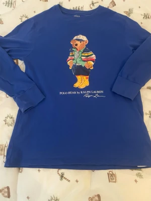 Blå långärmad Polo Bear t-shirt - Snygg blå långärmad t-shirt från Ralph Lauren med ikoniskt Polo Bear-tryck på bröstet. Tröjan har rund hals och är tillverkad i mjuk bomull. Perfekt för dig som gillar klassisk streetstyle med en twist.
