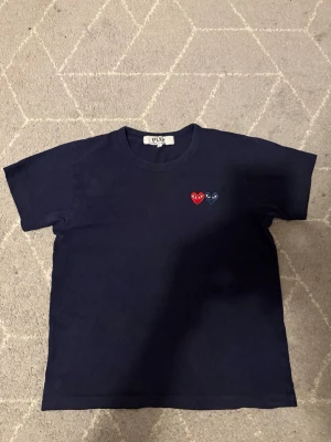 Mörkblå Comme des Garçons PLAY tee - Säljer en mörkblå t-shirt från Comme des Garçons PLAY med två broderade hjärtan på bröstet, ett rött och ett marinblått. Klassisk rund hals och korta ärmar. Perfekt för dig som gillar stilrena plagg med en twist. Det står L i den men sitter mer som en xs. Bara att fråga om ni undrar något😀