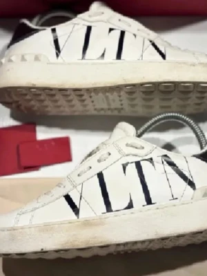 Vita VLTN sneakers från Valentino - Snygga vita sneakers från Valentino med stor svart VLTN-logga på sidan. Skorna har lågt skaft, vita snören och detaljer i svart vid hälen. Yttermaterialet är slätt läder och sulan har ett rutmönster för extra grepp.