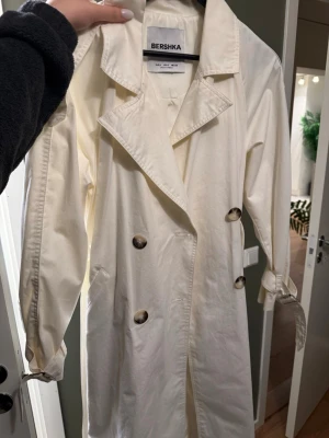 Vit trenchcoat från Bershka - Stilren vit trenchcoat från Bershka med klassisk krage, dubbelknäppning och stora knappar i brunmelerad plast. Jackan har bälte i midjan och spännen vid ärmsluten för extra detalj. Tillverkad i ett lätt tyg, perfekt för lager på lager speciellt till vår eller sommar, in princip oanvänd i storlek S