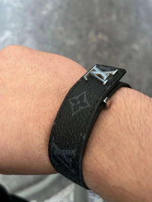 Louis Vuitton svart läderarmband - Snyggt svart armband från Louis Vuitton i präglat läder med det klassiska LV-märket i silverfärgad metall. Armbandet har diskreta monogramdetaljer och en stilren design som passar perfekt till din outfit.