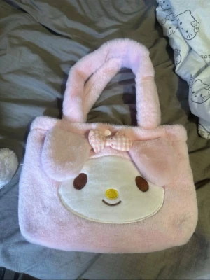 My Melody fluffig rosa handväska - Supersöt fluffig handväska i ljusrosa med My Melody-motiv från Sanrio. Väskan har mjuka handtag, ett broderat ansikte och en rosa-rutig rosett framtill. Perfekt för dig som älskar kawaii-stil och vill ha något gulligt till din outfit.