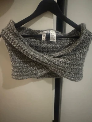 Grå stickad tubhalsduk H&M - Mysig grå stickad tubhalsduk från H&M med grov virkning och twistad design framtill. Perfekt för att hålla värmen under kyliga dagar och enkel att styla till olika outfits. Tillverkad i mjukt garn för extra komfort.