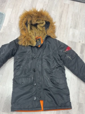 Svart parkas från Alpha Industries - Snygg svart parkas från Alpha Industries med fuskpäls på huvan och orange innerfoder. Jackan har flera fickor, dragkedja och knappar framtill samt den klassiska röda 'Remove Before Flight'-taggen på ärmen.