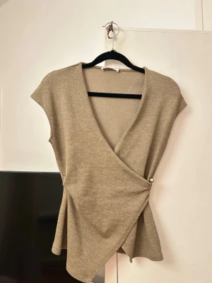 Beige omlottopp med knappdetalj - Snygg beige omlottopp i stickad kvalitet med v-ringning och korta ärmar. Toppen har en dekorativ knapp i sidan som håller omlotten på plats och ger en stilren look. Perfekt att matcha med jeans eller kjol för en trendig outfit.