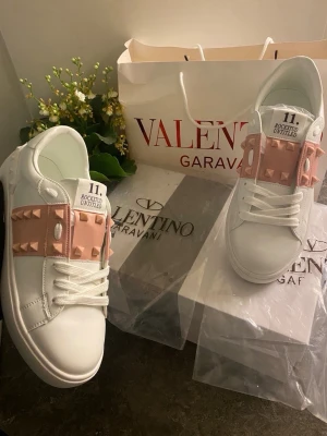 Valentino Garavani Skor - Snygga vita sneakers från Valentino med breda ljusrosa band över snörningen dekorerade med ikoniska nitar. Modellen heter Rockstud Untitled 11 och har en clean, modern look med coola detaljer. Perfekta för dig som vill sticka ut med exklusiva skor.
