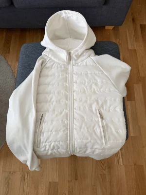 Vit Cardigan  - Säljer en vit Cardigan hoodie med quiltad front och rygg, dragkedja hela vägen och två fickor med dragkedja. Hoodien har en stor huva och ärmar i mjukt sweatshirtmaterial. Perfekt för dig som gillar en clean och sportig stil.