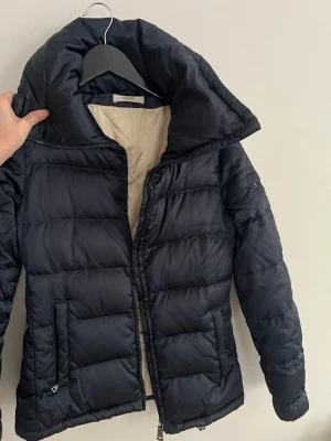 Prada Skidjacka - Blå pufferjacka från Prada med hög krage och dragkedja framtill. Jackan har också ett skärp inne. Skidjacka som är i storlek 40 men passar en S  Perfekt för kalla vinterdagar och ger en lyxig känsla med sin stilrena look.