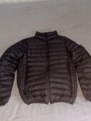 Svart dunjacka från Stone Island - Snygg svart dunjacka från Stone Island med klassisk patch på ärmen. Jackan har quiltad design, hög krage och dragkedja framtill. Perfekt för kalla dagar och ger en clean streetwear-look. Helt oskadad och nästan helt ny har använt i en vecka det är storlek s men passar M perfekt 