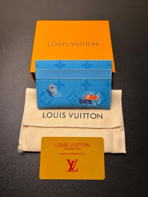 Blå Louis Vuitton korthållare - Snygg blå korthållare från Louis Vuitton med ikoniskt monogrammönster och färgglada illustrationer av bilar. Tillverkad i tåligt canvasmaterial och har flera fack för kort. Perfekt accessoar för dig som vill ha något unikt och trendigt i fickan.