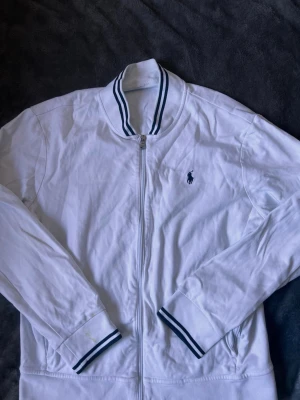  ziptröja från Polo Ralph Lauren - Klassisk vit ziptröja från Polo Ralph Lauren med blå logga på bröstet. Tröjan har ribbade muddar och krage med blå ränder samt dragkedja framtill. Tillverkad i mjukt bomullsmaterial och har en sportig, stilren look. Den har en fläck men går nog att få bort i tvätten men derför jag säljer för så billigt.
