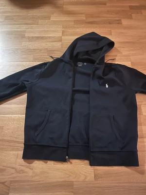 Navy blue Polo Ralph Lauren Zip Up - Snygg svart hoodie från Polo Ralph Lauren med dragkedja, två fickor framtill och klassisk vit logga på bröstet. Tillverkad i mjukt material och har en stor huva med dragsko. Perfekt för en chill och stilren look. Knappast använd