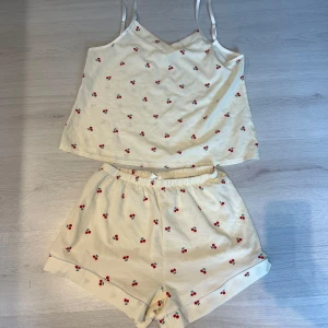 Pyjamasset med körsbär - Sött pyjamasset i mjukt material med fint körsbärsmönster 🍒 Består av linne och shorts.  Helt nytt och oanvänt. Perfekt som sommarpyjamas eller bekvämt mysset hemma.  📦 Skick: Ny / Oanvänd 👚 Storlek: M 🏷️ Märke: SHEIN 🎨 Färg: Offwhite med körsbär