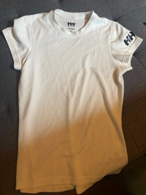 Vit t-shirt från Helly Hansen - Klassisk vit t-shirt från Helly Hansen med korta ärmar och rund hals. Diskret HH-logga på ärmen och i nacken. Tillverkad i mjuk bomull som känns skön mot huden. Perfekt basplagg till garderoben.