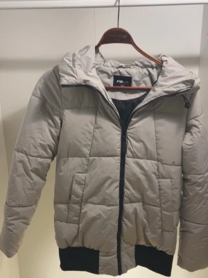 Beige pufferjacka från Fila - Snygg beige pufferjacka från Fila med svart dragkedja och svarta muddar. Jackan har en stor huva och quiltad design som ger en modern look. Perfekt för kalla vinterdagar och riktigt skön att bära. Materialet är syntet och håller värmen bra.