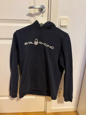 Mörkblå Sail Racing hoodie - Mörkblå hoodie från Sail Racing med vit logga tryckt på bröstet. Klassisk modell med huva och stor magficka framtill. Mjuk bomullskänsla och ribbade muddar vid ärmslut och nederkant. Perfekt för en avslappnad och sportig stil.