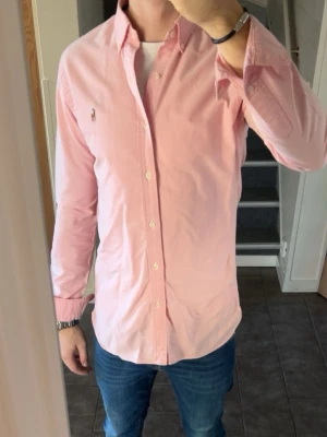 Skjorta Polo Ralph Lauren - Snygg rosa skjorta från Polo Ralph Lauren med klassisk button-down krage och broderad logga på bröstet. Skjortan har lång ärm, normal passform och är tillverkad i mjuk bomull. Perfekt för en stilren och avslappnad look. Utmärkt skick utan tecken på användning. Storlek small med slim fit. 