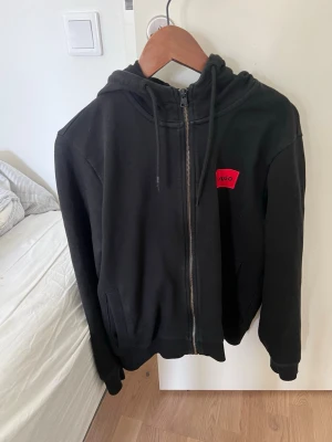 Svart zip hoodie från Hugo - Snygg svart hoodie med dragkedja från Hugo. Hoodien har en huva med dragsko och en röd logopatch på bröstet. Tillverkad i mjukt material och har ribbade muddar vid ärmslut och nederkant. Perfekt för en avslappnad och stilren look. Tröjan säljs för 1350kr ny 